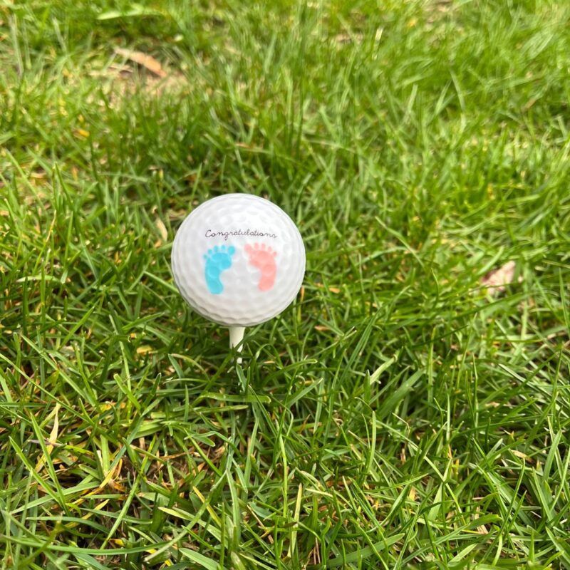 Custom golf ball printed with baby footprints and the word Congratulations, resting on a tee in the grass