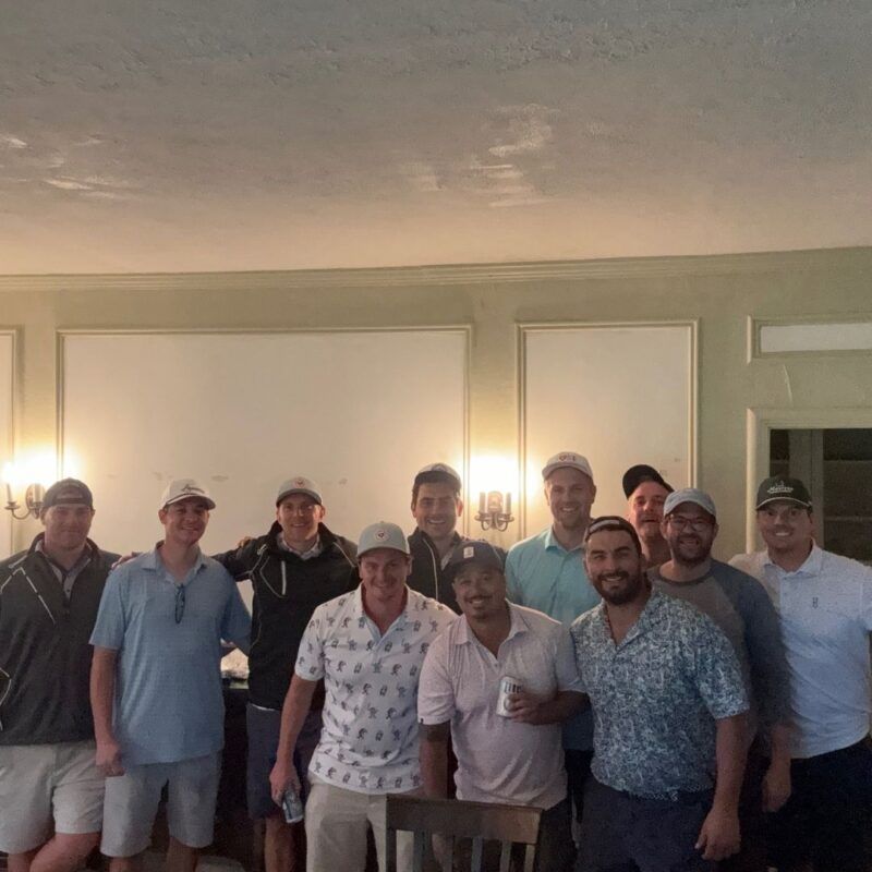 Group of about a dozen men in golf caps gathered inside a clubhouse for a group photo after a round